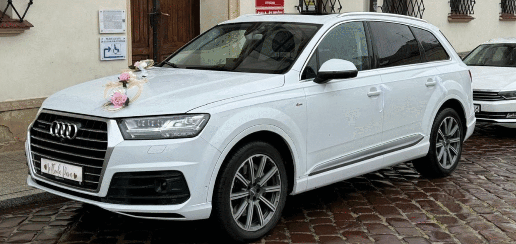 Samochód marki Audi, model Q7. Samochód koloru białego, na masce ma kremowo różowe przystrojeni weselne. Zdjęcie przedstawia samochód z przodu oraz lewej strony.