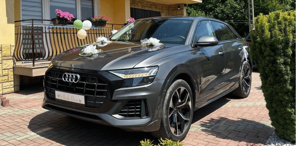 Samochód marki Audi model Q8. Zdjęcie zostało zrobione frontowej oraz lewej strony samochodu. Samochód jest ciemny szary przystrojony w białe dekorację weselne.