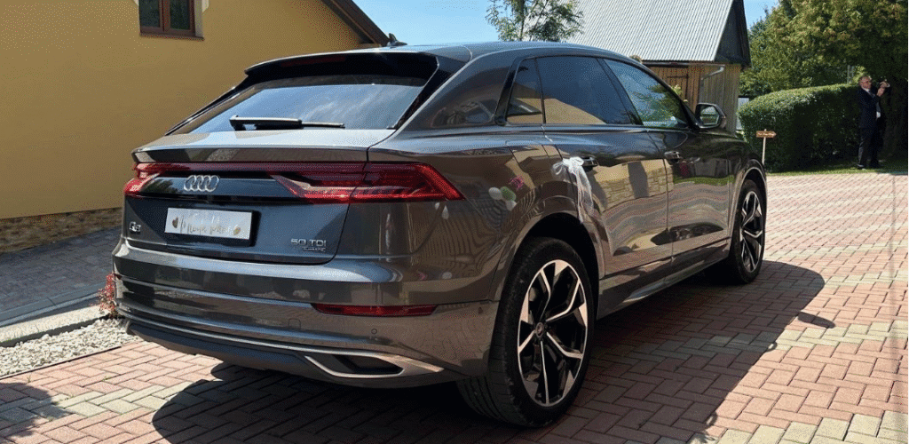 Samochód marki Audi, model Q8. Jest to zdjęcie tyłu samochodu oraz prawej strony. Samochód jest koloru czarnego i jest przyzdobiony w białe dekoracje weselne.