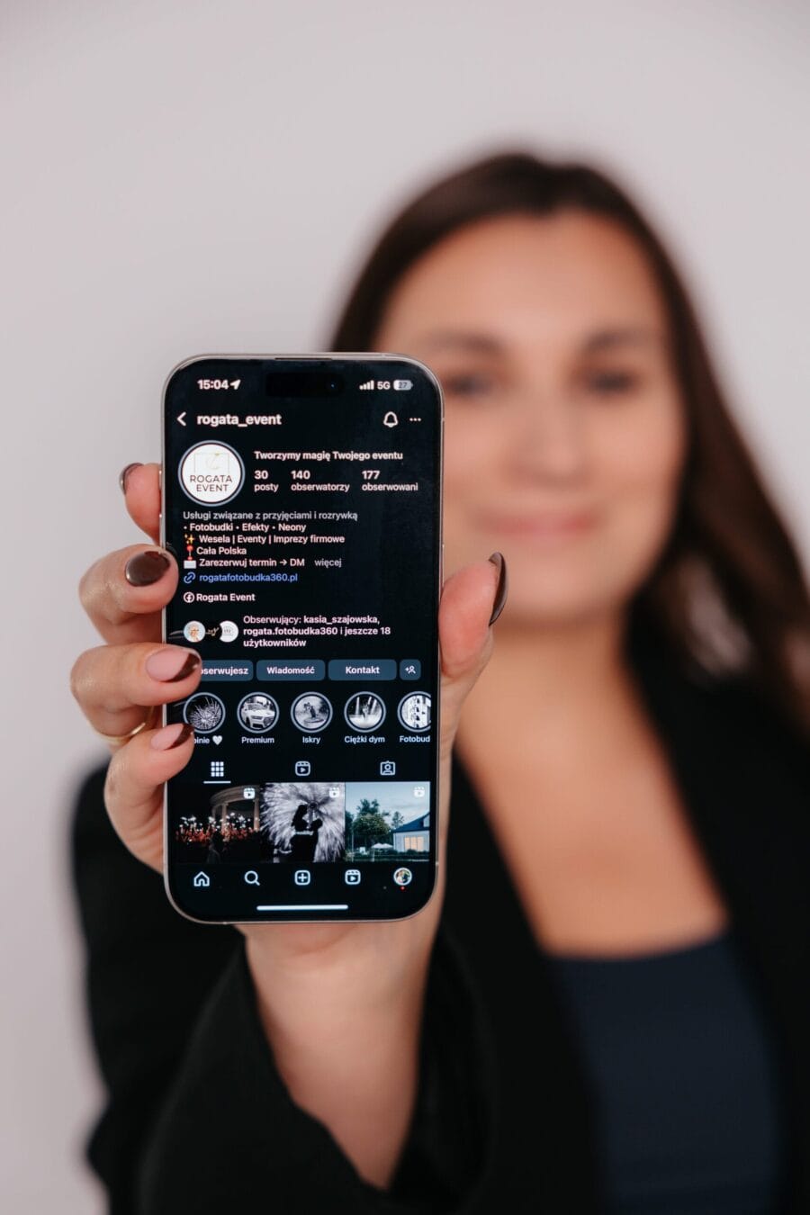 Zdjęcie przedstawia zamazaną Kobietę, która trzyma ręku telefon, jest on wyostrzony na zdjęciu. Na ekranie telefonu jest wyświetlona strona social media firmy Rogata Event