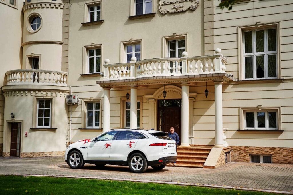 Zdjęcie przedstawia samochód marki Jaguar, model F-Pace. Jest koloru białego przyzdobionego w czerwone dekoracje weselne.