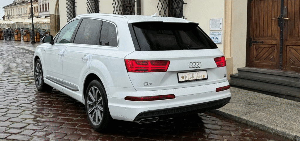 Samochód marki Audi, model Q7. Samochód jest koloru białego, jest to zdjęcie przedstawiające tył oraz lewą stronę samochodu.